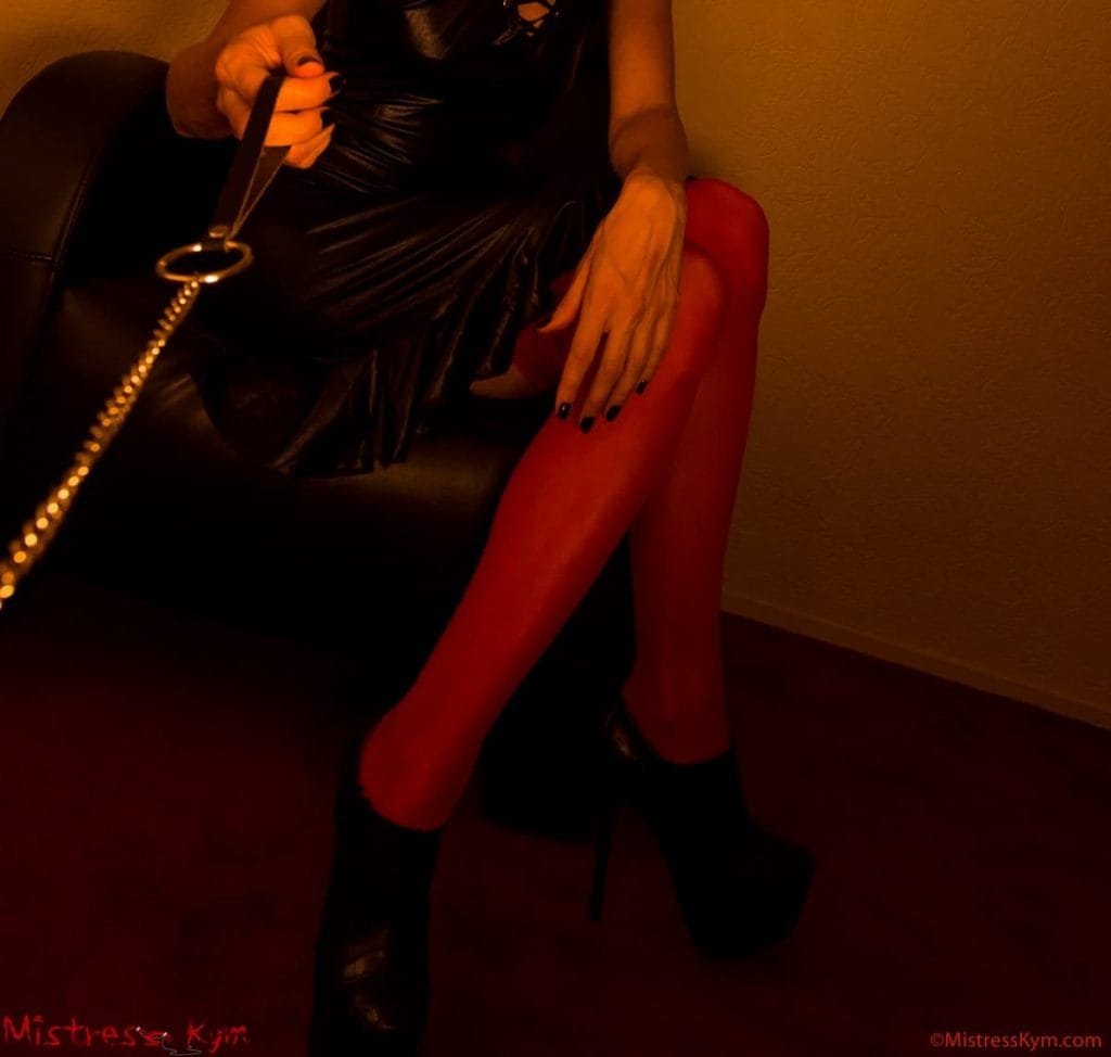 Maîtresse Kym en bas et leggings rouges tenant son esclave au collet et en laisse.