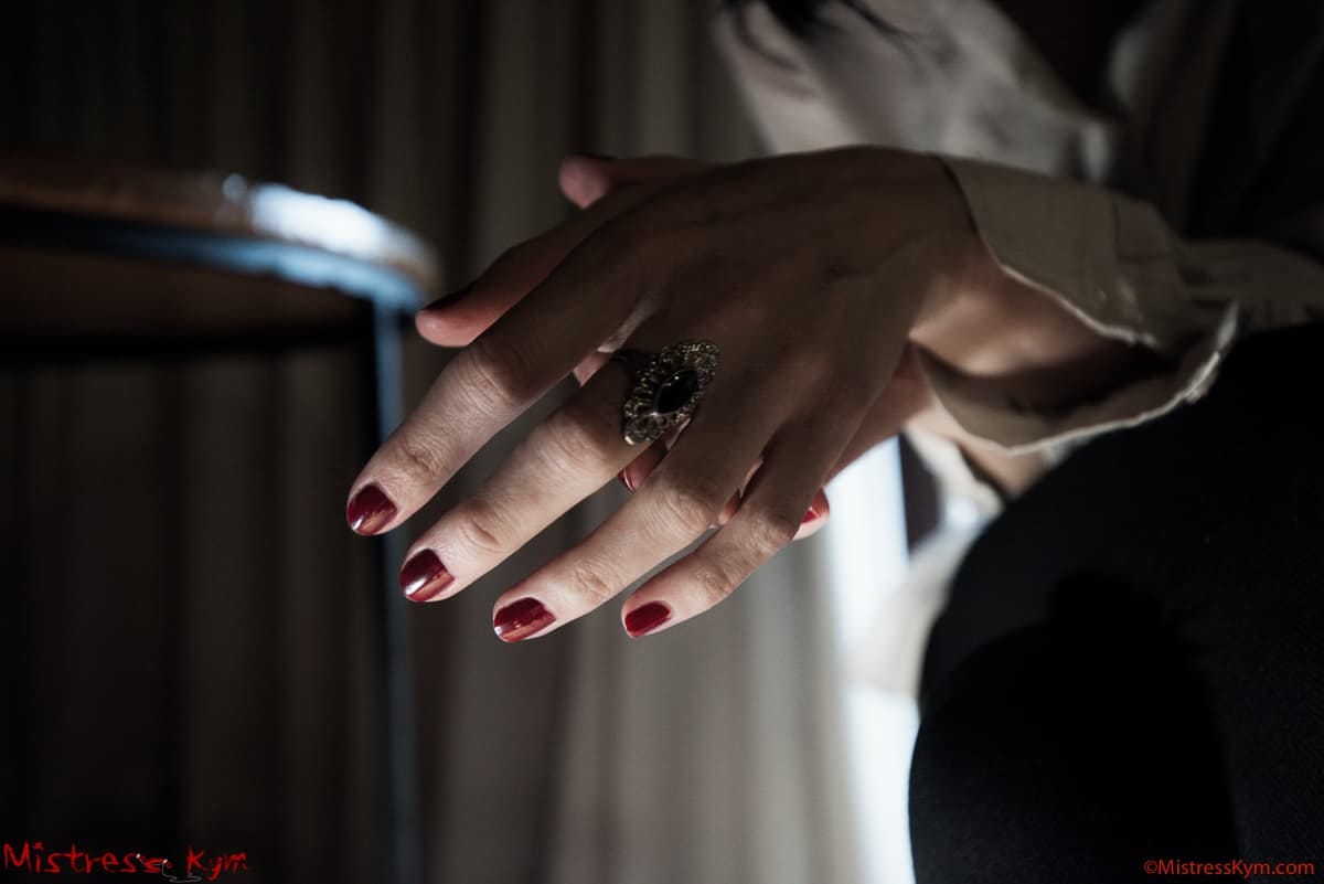 Mistress Kym visar sina veneriska händer med rödpolerade naglar och sin ring.
