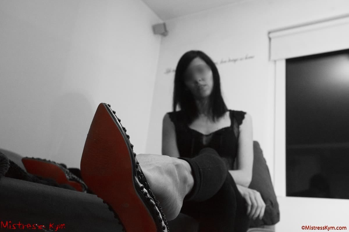Stăpâna Kym vă arată talpa roșie a pantofilor ei negri cu crampoane și picioarele ei lungi.