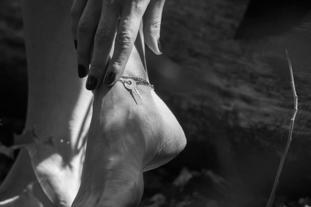Mistress Kym está segurando as chaves do seu cinto de castidade no tornozelo dela com os pés descalços.